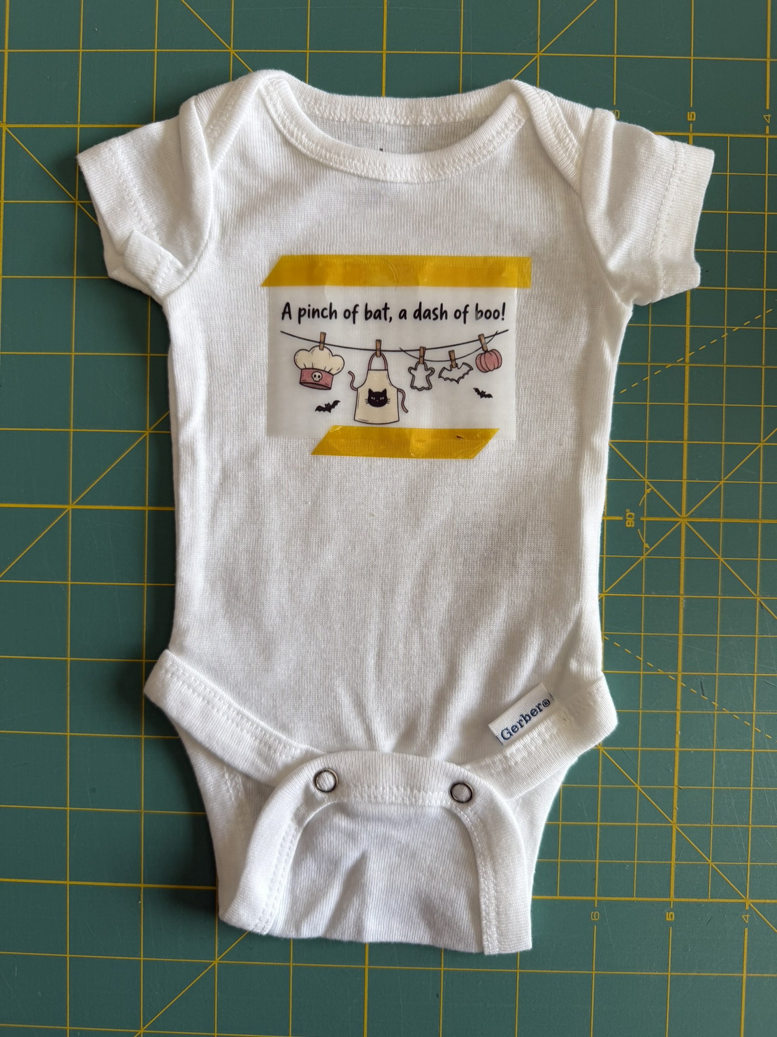 White preemie bodysuit with a DTF transfer Halloween graphic taped to the front so that it can be pressed onto the fabric. The oneisie is laid out on a green cutting mat that has a yellow grid pattern.