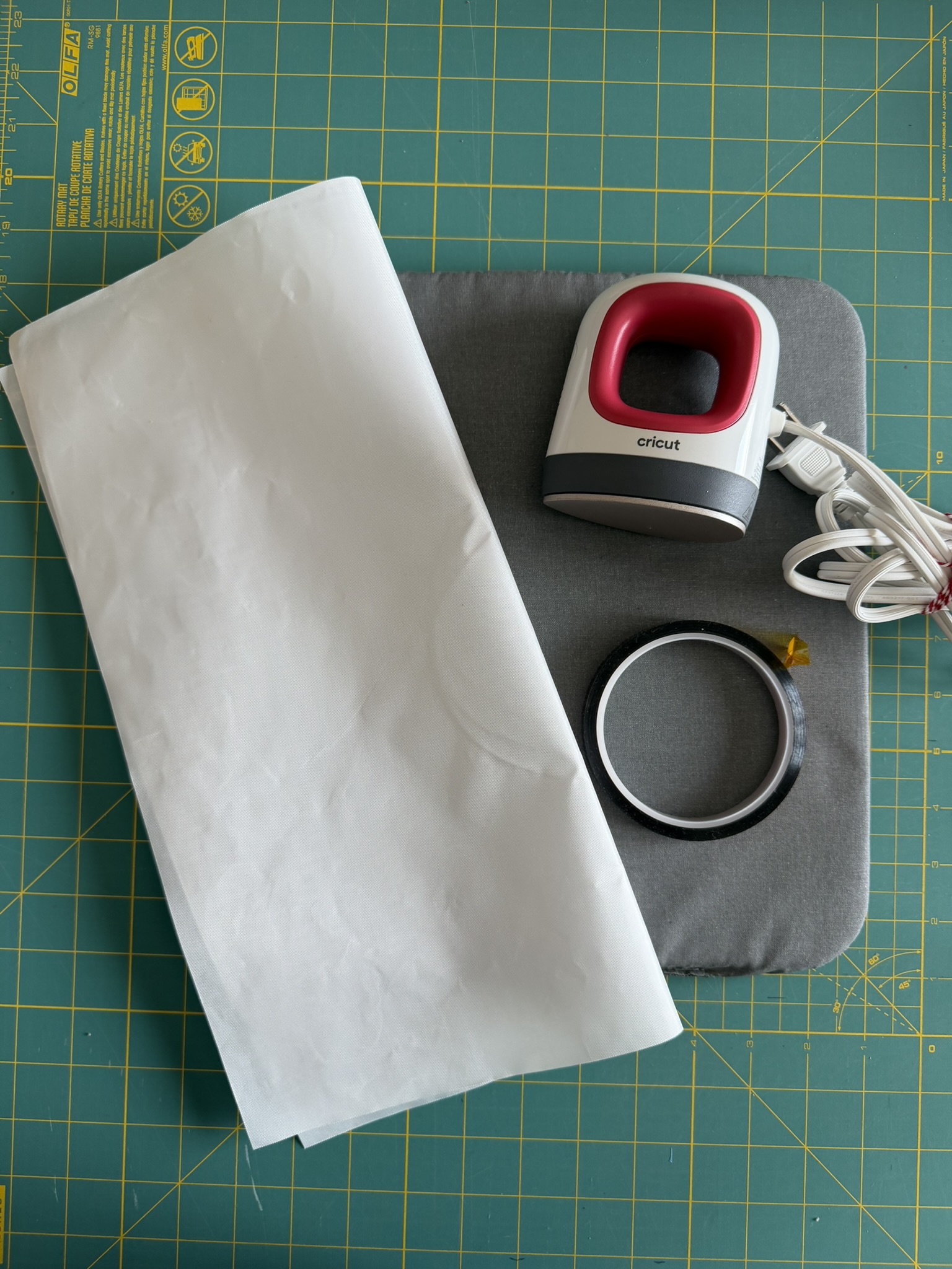 A teflon pressing sheet, Cricut mini heat press, heat-resistant pressing mat and heat tape placed on a cutting mat to showcase all the tools needed for a DIY Halloween preemie bodysuit.