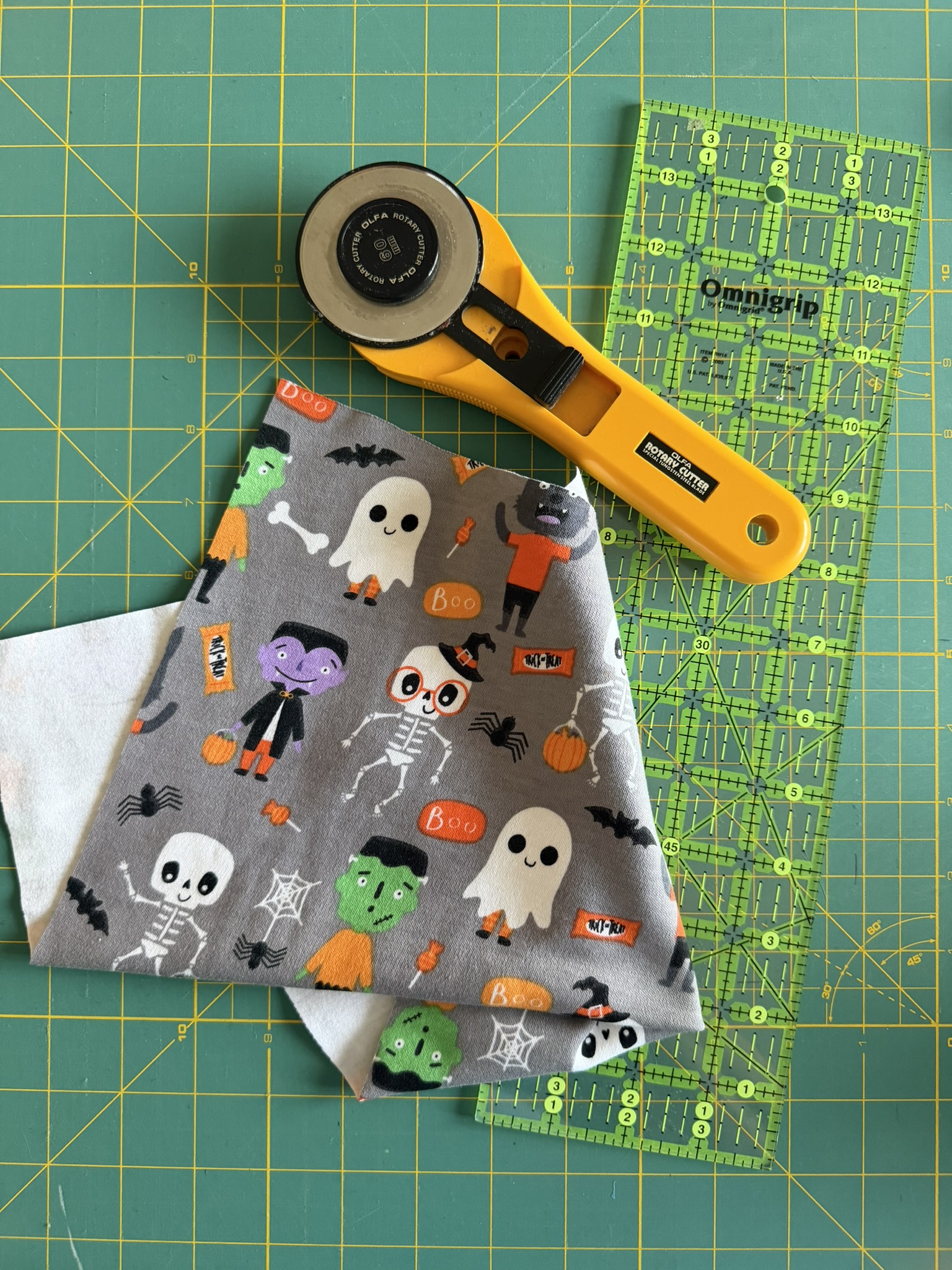Knit fabric with Halloween-themed graphics next to a yellow rotary cutter, Omnigrid ruler on a green cutting mat.