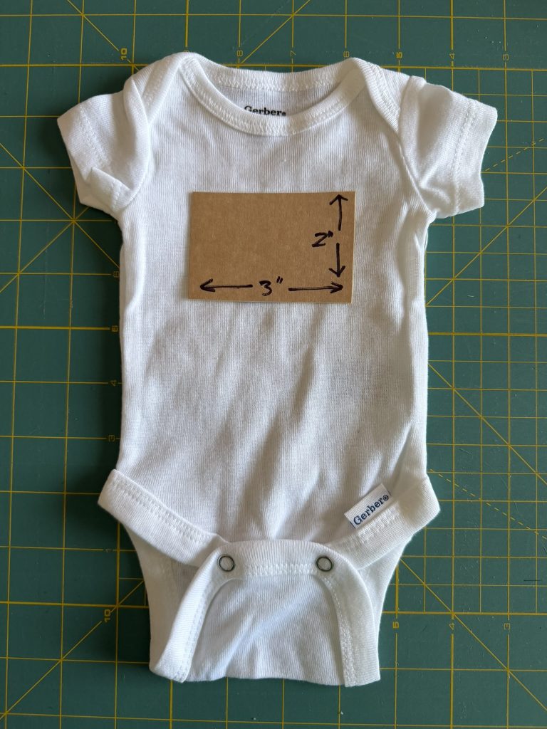 White preemie bodysuit on a green cutting mat with a 3 inch by 2 inch cardboard cut out on the front of the bodysuit to showcase what that size looks like on the fabric.