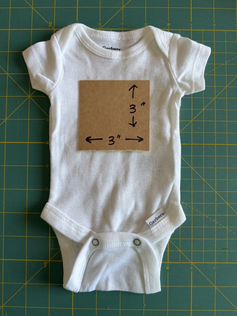 White preemie bodysuit on a green cutting mat with a 3 inch by 3 inch cardboard cut out on the front of the bodysuit to showcase what that size looks like on the fabric.