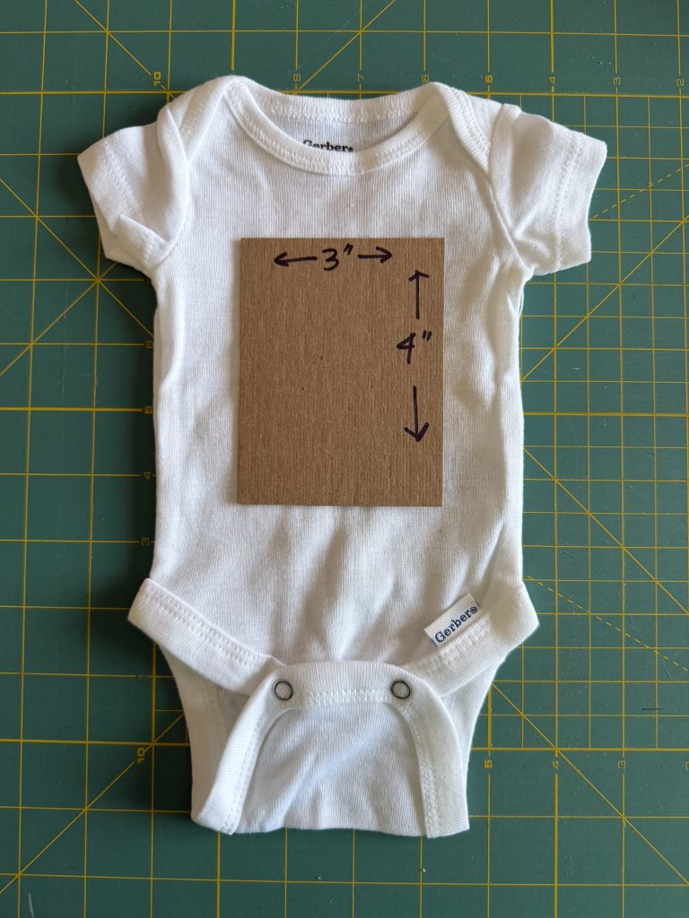 White preemie bodysuit on a green cutting mat with a 3 inch by 4 inch cardboard cut out on the front of the bodysuit to showcase what that size looks like on the fabric.