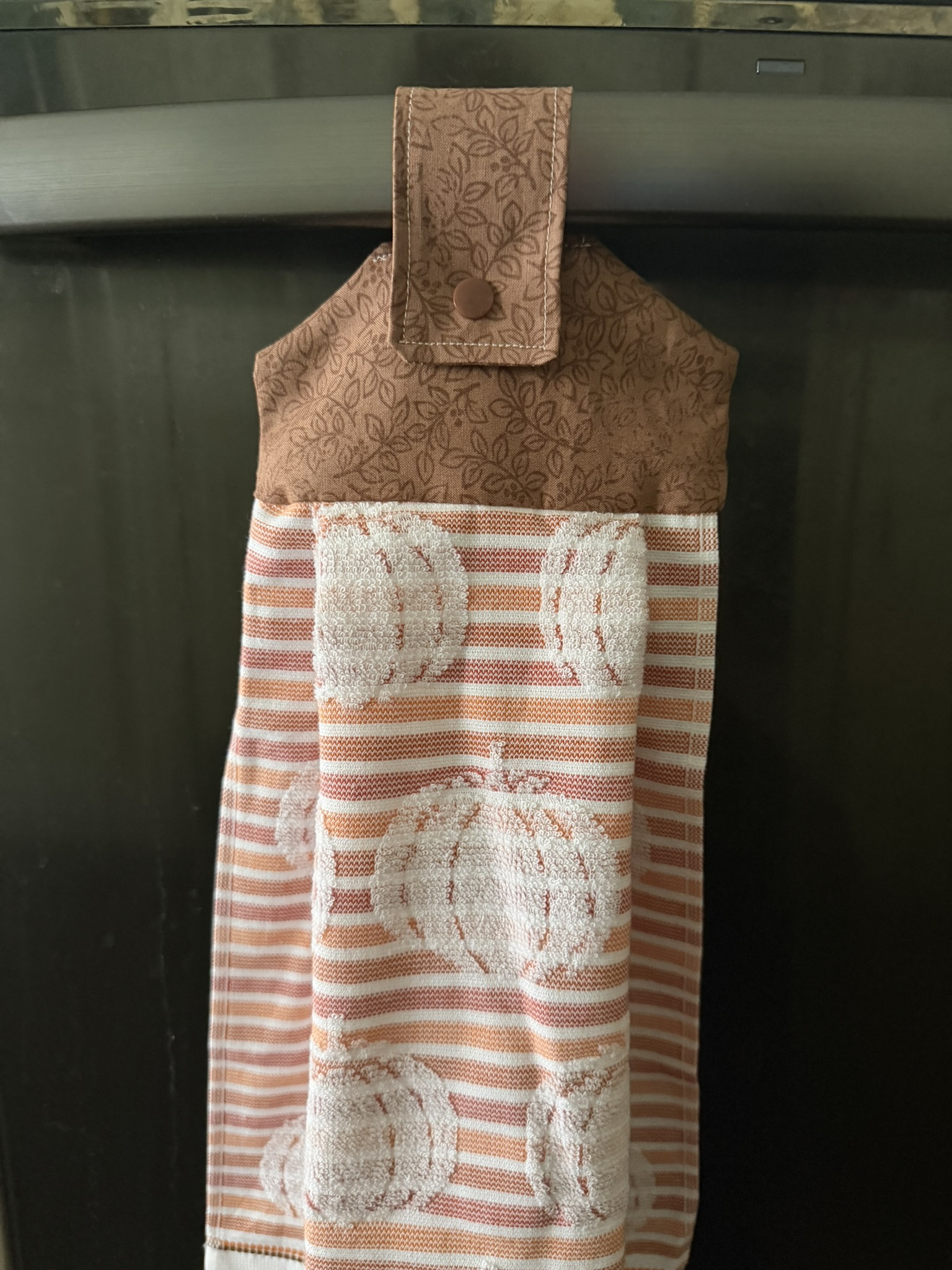 Fall-themed hanging dish towel hanging from the handle of a dishwasher.