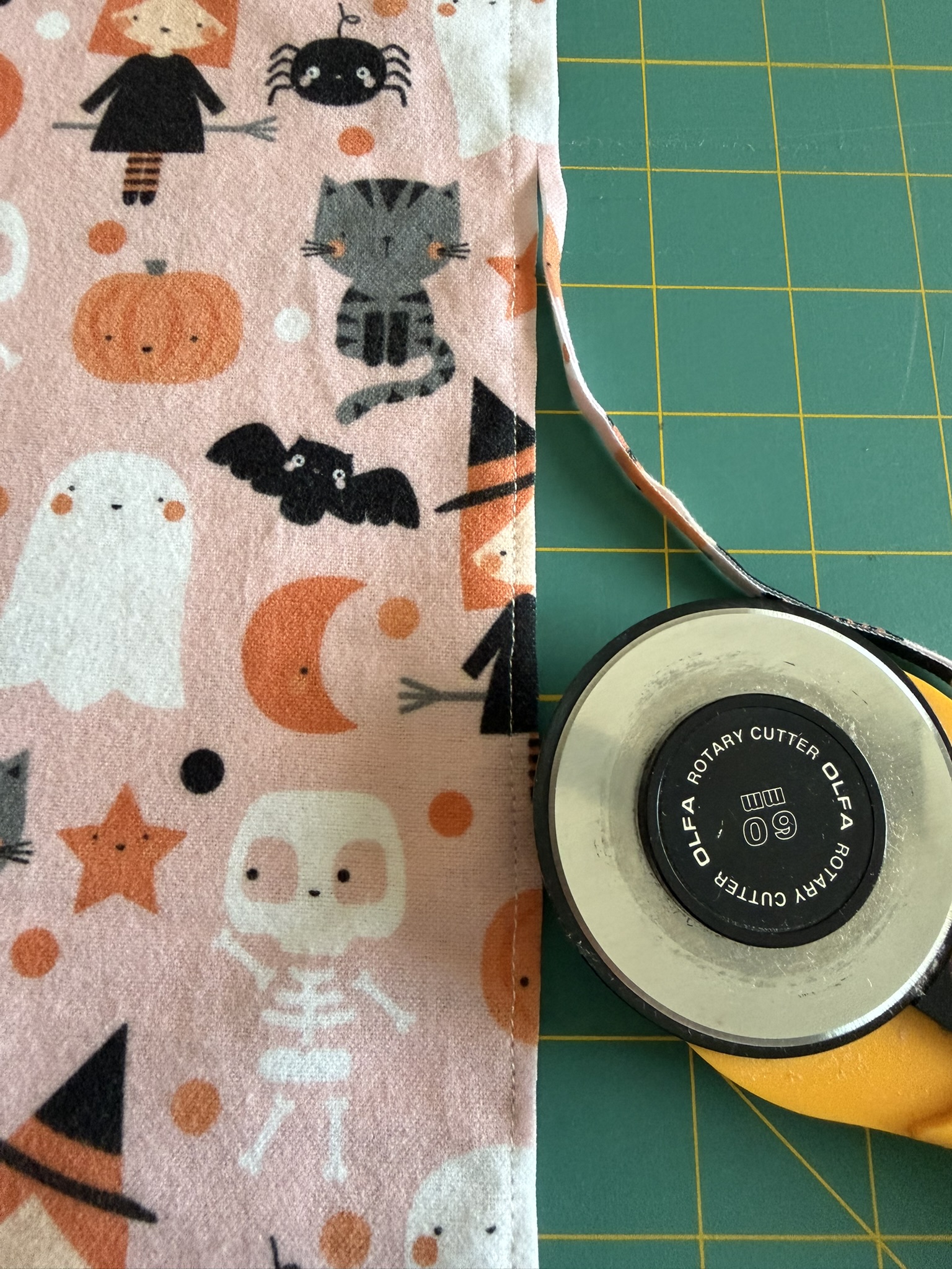 Halloween-themed fabric that is being trimmed by a yellow-handled rotary cutter on a green cutting mat.